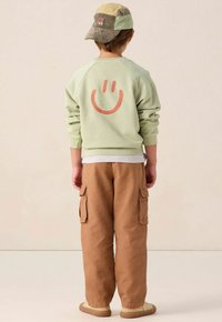 Sweatshirt vert avec un grand visage souriant orange dans le dos, associé à un pantalon cargo beige avec des poches latérales et des chaussures beiges.