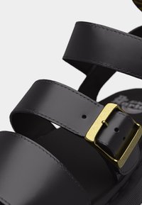 Λουράκια μαύρου δερμάτινου σανδάλιου με λεπτομερή ραφές και χρυσό μεταλλικό ρυθμιζόμενο αγκράφα, σε κοντινό πλάνο.