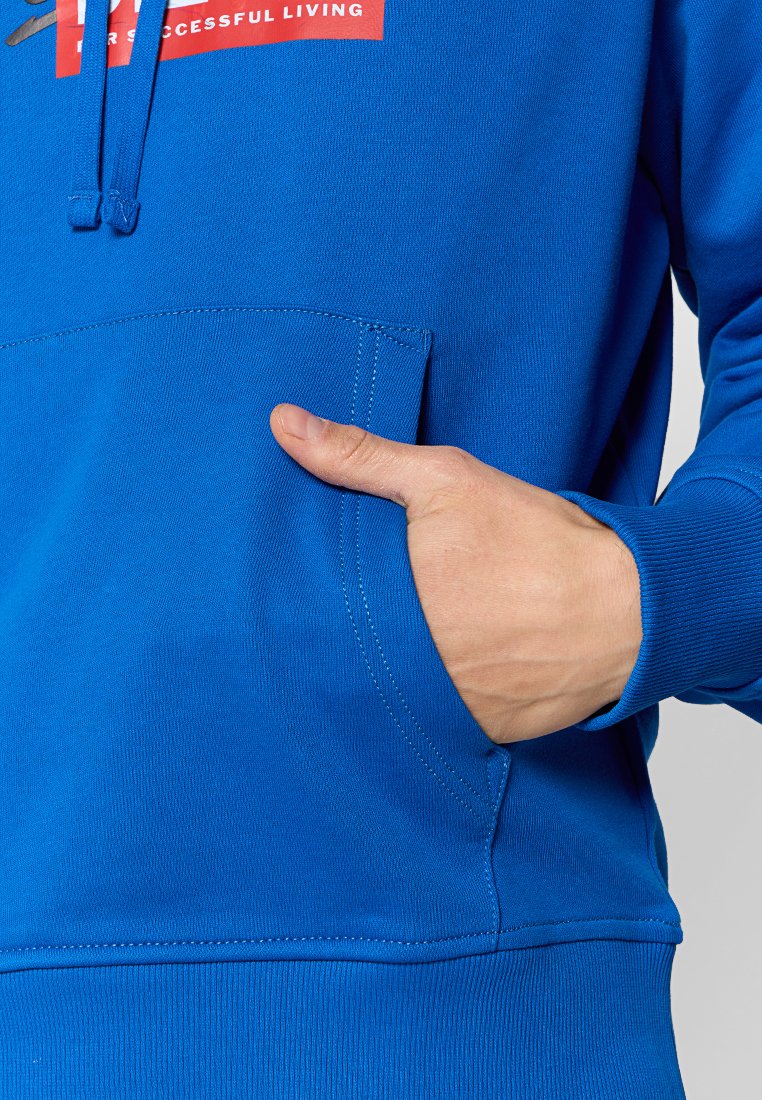 Sudadera azul hecha de tejido suave, con un bolsillo canguro en la parte delantera y un logo rojo en el pecho. Se pueden ver detalles de costura a lo largo de los bordes.