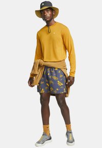Long-sleeve mustard shirt, navy shorts with yellow insect patterns, beige sweater tied at the waist, black bucket hat, gray sneakers with accents.