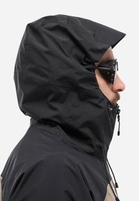 Veste à capuche noire avec une surface texturée, des cordons réglables et un design épuré. Présente un panneau contrastant beige clair à l'épaule.