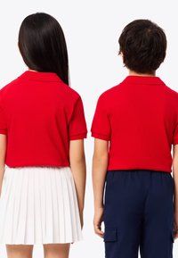Röda pikétröjor med korta ärmar och krage bärs av ett barn och en ung person, kombinerade med en vit veckad kjol och marinblå cargobyxor.