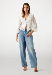 Witte blouse met lange mouwen, versierd met bloemenborduursels en een striksluiting, gecombineerd met lichtblauwe denim jeans met wijde pijpen en bruine sandalen.