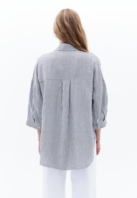 Camicia a righe con linee verticali bianche e blu navy, vestibilità comoda, maniche arrotolate e dettaglio sul retro. Il tessuto sembra leggero e traspirante.