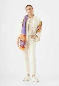 Veste en denim clair sur un top beige et un pantalon assorti, avec une écharpe à motifs colorés drapée sur une épaule et des baskets blanches.