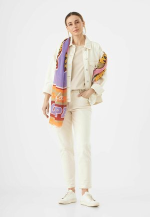Veste en denim clair sur un top beige et un pantalon assorti, avec une écharpe à motifs colorés drapée sur une épaule et des baskets blanches.