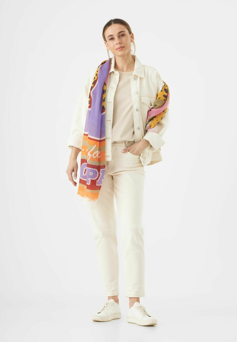 Veste en denim clair sur un top beige et un pantalon assorti, avec une écharpe à motifs colorés drapée sur une épaule et des baskets blanches.