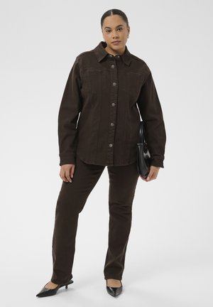 Frau trägt eine dunkelbraune, geknöpfte Jacke und passende Hose, hält eine schwarze Handtasche und steht vor einem hellen Hintergrund.