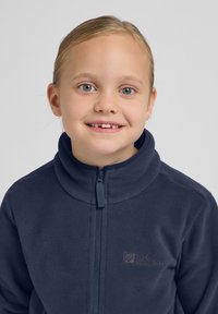 Tumesinise fliisjakiga, millel on kõrge kaelus ja eespool tõmblukk, ning millel on rinnale tikitud väike logo. Pehme tekstuur.