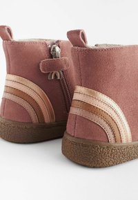 Cizme ankle din suede roz, cu căptușeală din blană de oaie, având un fermoar pe lateral și talpă din cauciuc maro. Împodobite cu dungi din piele curbate în nuanțe variate.