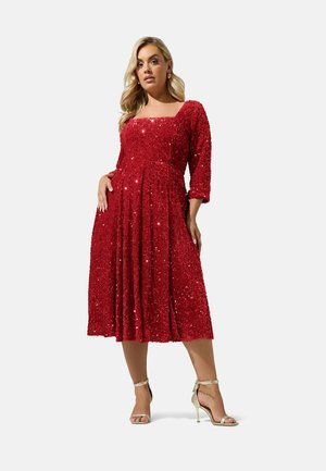 Mujer con vestido midi rojo de lentejuelas, con manga tres cuartos y escote cuadrado, combinado con sandalias de tacón alto beige.