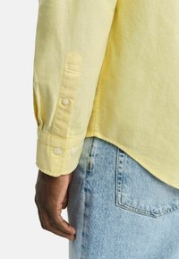 Geel langemouw overhemd met knoopmanchetten, zachte katoenen textuur en een gebogen onderrand, gecombineerd met lichtblauwe denim jeans.