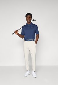 Jūrinio stiliaus polo marškiniai su horizontaliomis tekstūromis, baltas Nike logotipas, šviesiai smėlin�ės spalvos kelnės ir baltos golfo bateliai. Golfo lazda prispausta prie peties.