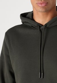 Felpa con cappuccio verde scuro con una texture morbida, cappuccio con coulisse e punte in metallo, design semplice e minimalista. Nessuna tasca o logo visibile.