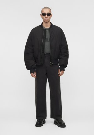 Homme portant une veste bomber noire oversize, pantalon noir ample, chaussures noires épaisses, lunettes de soleil, debout devant un fond blanc uni.