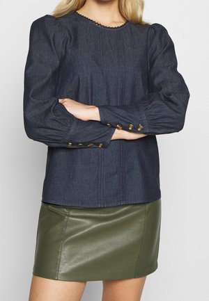 Vrouw draagt een donkerblauwe blouse met lange mouwen en gouden knopen op de manchetten, samen met een olijfgroene leren minirok, armen over elkaar op de taille.