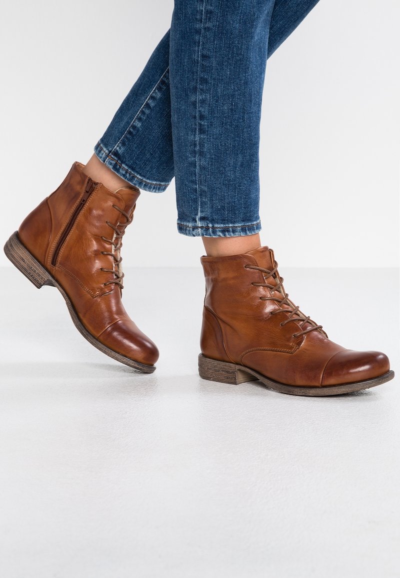 Bottes en cuir marron à lacets et fermeture éclair latérale, portées avec un jean en denim bleu, sur une personne debout sur un sol blanc.