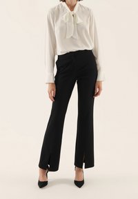 Bílá blůza s výstřihem ve tvaru mašle a dlouhými rukávy, kombinovaná s černými kalhotami s vysokým pasem a bočními rozparky, doplněná černými lodičkami.