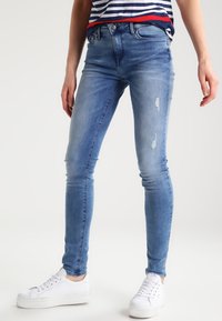Jeans ajustados en denim azul claro con desgastes y parches rasgados, que presentan un clásico diseño de cinco bolsillos y trabillas para cinturón.