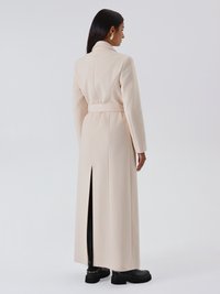 Cappotto lungo beige con vita con cintura, colletto con risvolto e spacco posteriore. Tessuto liscio, vestibilità sagomata, indossato sopra pantaloni neri e stivali robusti.