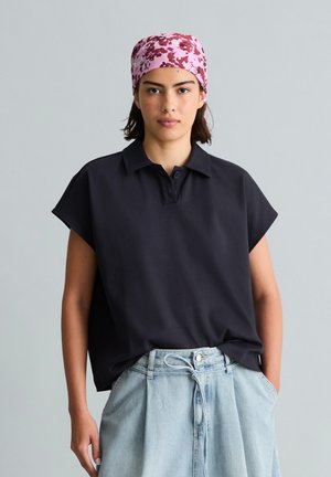 Femme portant un foulard rose à motifs floraux, un t-shirt noir ample à manches courtes et un jean taille haute bleu clair, les mains dans les poches.
