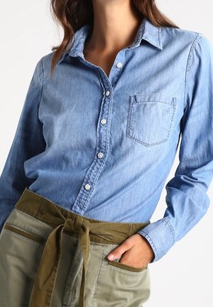 Kvinde iført en lyseblå denim skjorte med knapper og brystlomme samt olivengrønne bukser bundet i taljen med et bælte i samme stof.