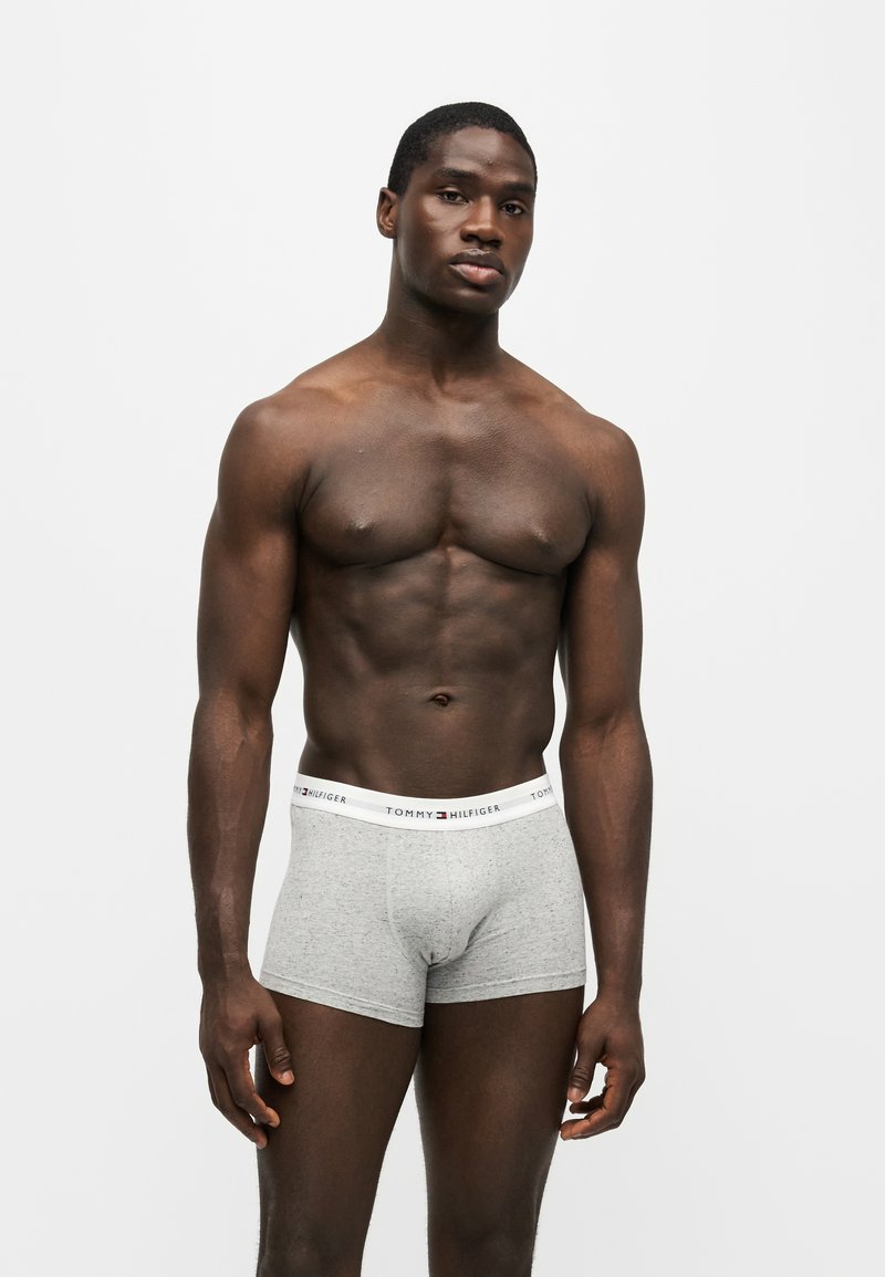 Atletisk mørkhudet mand står mod hvid baggrund iført lysegrå Tommy Hilfiger boxershorts med logo på linningen.