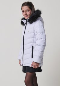Chaqueta de plumas blanca con capucha forrada de piel negra, cierre frontal con cremallera y bolsillos laterales. Presenta un pequeño logo de corazón en el pecho.