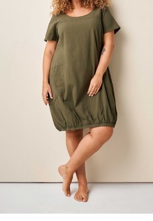 Woman with curly hair wearing a loose olive green short-sleeve dress standing barefoot against a plain beige wall.