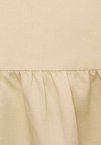 Tessuto beige con una trama testurizzata, caratterizzato da un orlo arricciato sulla parte inferiore. Il materiale appare morbido e leggero.