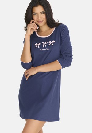 Frau trägt ein navyblaues langärmliges Kleid mit pinken Schleifenmustern und dem Text „c'est très bien“, lächelt und posiert mit der Hand nahe am Gesicht.