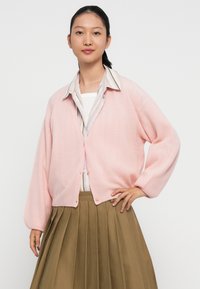 Cardigan a maglia di un rosa chiaro con una finitura testurizzata, chiusura con bottoni sul davanti e maniche ampie, indossato sopra una camicia con colletto e una gonna plissettata beige.