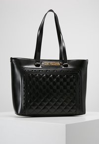 Black leather tote bag featuring a quilted design on the front, twin handles, and a gold-tone logo plaque at the top.