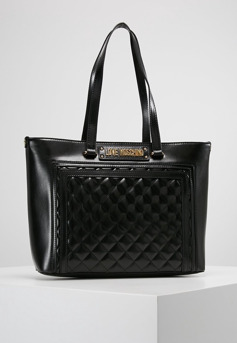 Black leather tote bag featuring a quilted design on the front, twin handles, and a gold-tone logo plaque at the top.