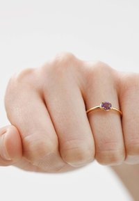 Anillo de oro con una gema circular morada, engastada en seis garfios, presentado sobre una mano texturizada contra un fondo claro.