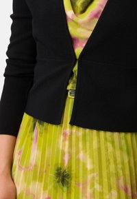 Černý zkrácený kardigan s texturovaným povrchem, nošený přes plisované šaty s zeleným základem a růžovým květinovým vzorem.