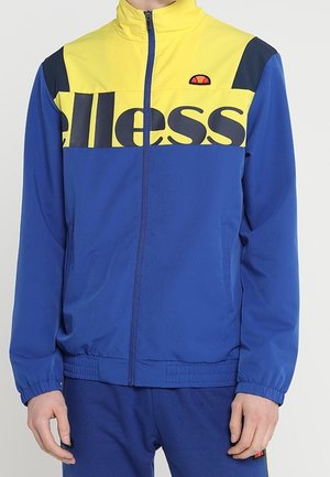 Homme portant une veste de sport zippée bleue et jaune avec le logo "ellesse" sur la poitrine, manches et taille élastiques.