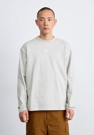 Tricou deschis gri cu mânecă lungă, fabricat din bumbac, având un mic logo alb Nike pe piept. Asortat cu pantaloni cargo maro.