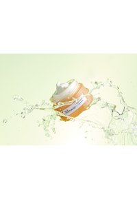 Oranges Glas mit Gesichtsfeuchtigkeitscreme und weißem Deckel, das durch Wasser spritzt, mit einem Etikett, auf dem "Gesichtsfeuchtigkeitscreme" und Inhaltsstoffe stehen.