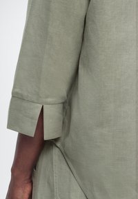 Λεπτομέρεια μανικιού ανοιχτόχρωμου ελαιόχρωμου πουκάμισου από λινό με σχίσιμο στην μανσέτα, σε σκούρο δέρμα χέρι, που δείχνει την υφή του υφάσματος και τη ραφή.