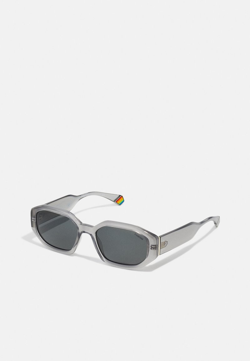 Gafas de sol grises transparentes con lentes hexagonales, tintados de gris oscuro, que presentan un pequeño acento de arcoíris en la patilla y herrajes de plata pulida.