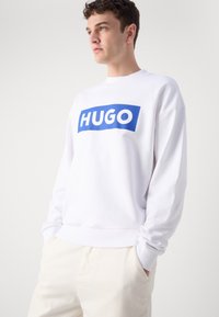 Balta džemperis, pagamintas iš minkštos medžiagos, su dideliu mėlynu stačiakampiu logotipu, ant kurio yra užrašas "HUGO" ryškiai baltomis raidėmis, laisvo kirpimo dizainas.