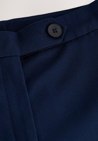 Primo piano di tessuto blu scuro che mostra una cintura con un bottone nero a quattro fori fissato su una linguetta di tessuto.