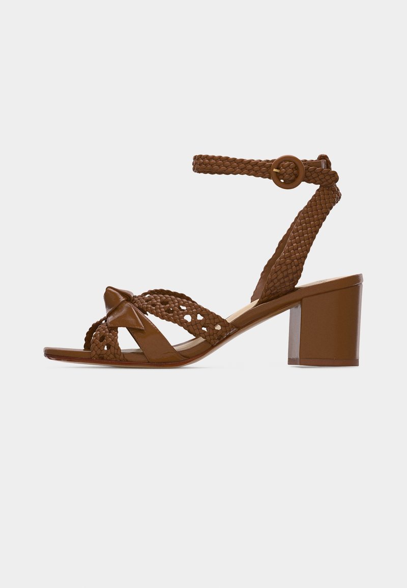 Καφέ δερμάτινο σανδάλι με τακούνι, με πλεγμένα λουράκια, αγκράφα στον αστράγαλο και χοντρό τακούνι, απεικονισμένο πλαγίως σε λευκό φόντο.