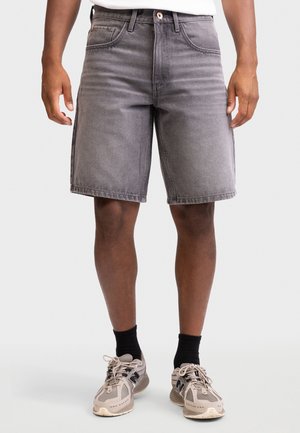 Mężczyzna w średniej długości wyblakłych szarych szortach dżinsowych, czarnych skarpetach i beżowych tenisówkach stoi na jednolitym tle.