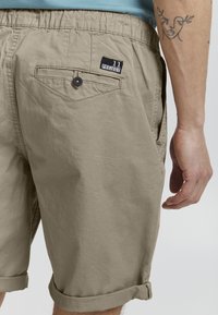 Beige shorts lavet af let materiale med en elastisk talje, opfoldede manchetter og en baglomme med et sort mærke.