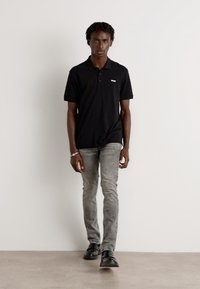 Polo negro de manga corta, jeans ajustados grises y zapatos negros. El modelo tiene el cabello de longitud media y está de pie contra un fondo simple.