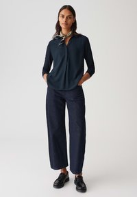 Marineblouse met driekwart mouwen, knoopdetails, gecombineerd met wijde, donkere denimbroek en zwarte veterschoenen. Groene sjaal als accent.