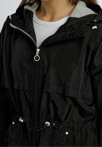 Schwarze Windjacke mit durchgehendem Reißverschluss, grauem Kapuzenfutter, Tunnelzug in der Taille und zwei Seitentaschen. Glattes, leichtes Material ohne Muster.