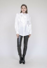 Blouse en satin blanc à manches longues et col à nouer, assortie à des leggings en cuir noir et des bottines. Texture lisse avec une finition brillante.